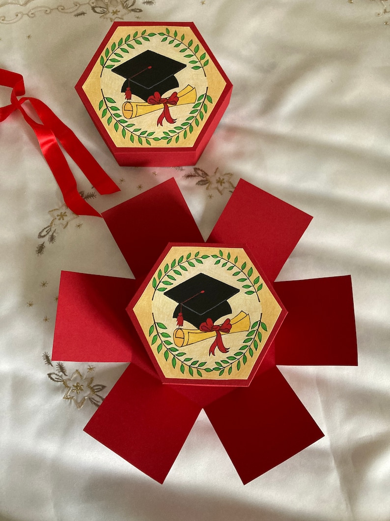 May include: A red hexagonal box with a graduation cap and diploma design. The box is open and the sides are folded out to reveal the interior.