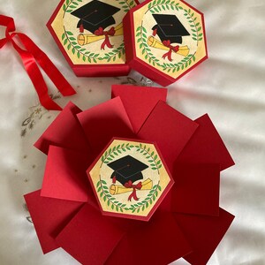 May include: Red explosion box with a graduation cap and diploma design. The box is closed and has a hexagon shape. There are two smaller hexagon boxes with the same design.