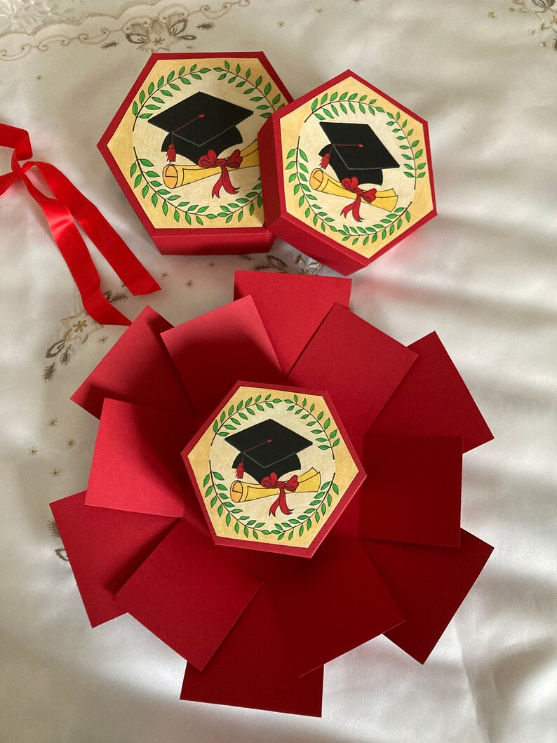 May include: Red explosion box with a graduation cap and diploma design. The box is closed and has a hexagon shape. There are two smaller hexagon boxes with the same design.