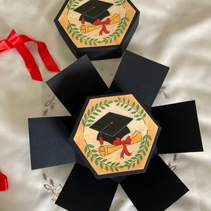May include: A black and gold graduation themed explosion box with a black graduation cap and a red ribbon on a diploma scroll. The box is open and has six sides.