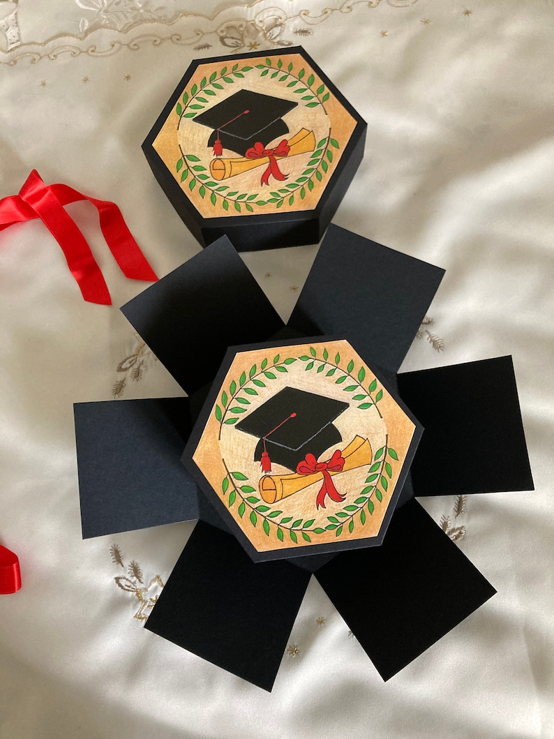 May include: A black and gold graduation themed explosion box with a black graduation cap and a red ribbon on a diploma scroll. The box is open and has six sides.