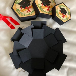 May include: A black explosion box with a graduation cap and diploma design. The box is made of cardstock and has a hexagonal shape. There are three smaller boxes with the same design.