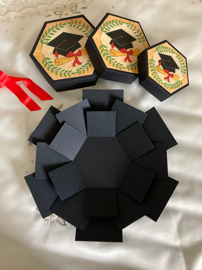 May include: A black explosion box with a graduation cap and diploma design. The box is made of cardstock and has a hexagonal shape. There are three smaller boxes with the same design.