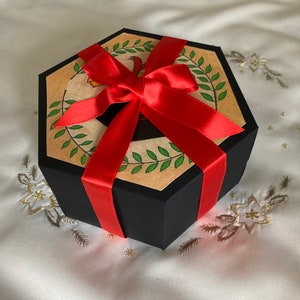 May include: A hexagonal gift box with a black lid and a red ribbon. The box is decorated with a brown paper with a green leaf design.