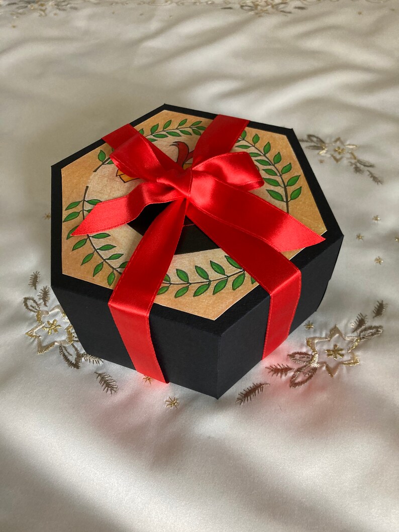 May include: A hexagonal gift box with a black lid and a red ribbon. The box is decorated with a brown paper with a green leaf design.