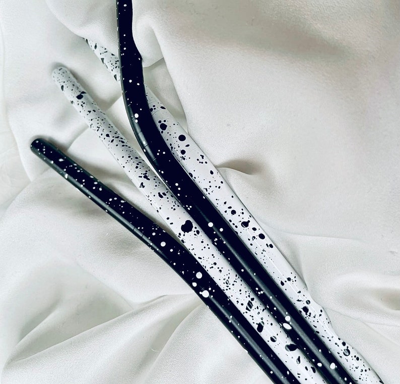 May include: Four slender, curved metal straws with a speckled black and white design. The straws are arranged diagonally against a white background, creating a visually appealing contrast. The design features a pattern of black and white spots.