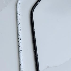 May include: Two reusable metal straws. One is white with black speckles, the other is black with white speckles. Both straws have a curved design. The straws are designed for drinking beverages.