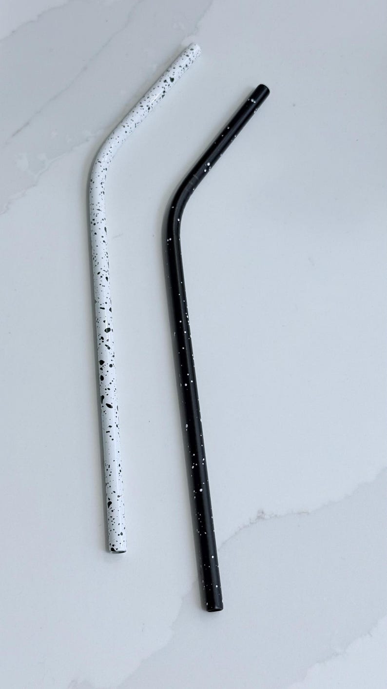 May include: Two reusable metal straws. One is white with black speckles, the other is black with white speckles. Both straws have a curved design. The straws are designed for drinking beverages.