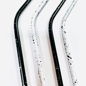 May include: Four stainless steel straws with a curved design. Two straws are black with white speckles, and two are white with black speckles. The straws are angled and appear to be reusable.