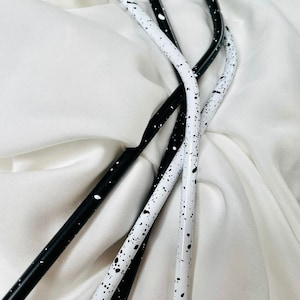 May include: Four reusable metal straws. Two are white with black speckles, and two are black with white speckles. The straws are curved and rest on a white, textured fabric.