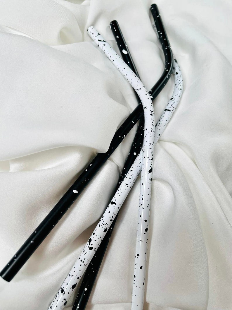 May include: Four reusable metal straws. Two are white with black speckles, and two are black with white speckles. The straws are curved and rest on a white, textured fabric.