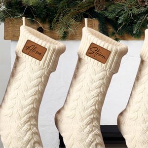 May include: Off-white cable knit Christmas stockings featuring a folded cuff with a rectangular brown leather patch. The patch on the left stocking is engraved with the name Alison, and the patch on the right stocking is engraved with the name Steven.