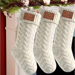 May include: Three white knitted Christmas stockings with brown leather name tags. The names on the tags are "Amanda", "Shawn", and "Becky".