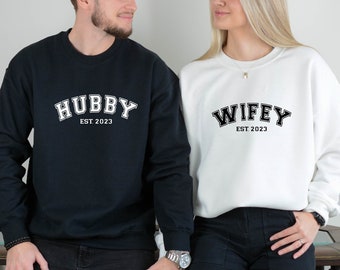 Peut inclure: Un sweat-shirt noir avec le texte "Hubby Est. 2023" et un sweat-shirt blanc avec le texte "Wifey Est. 2023".