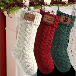 May include: Three knitted Christmas stockings with personalised leather name tags. The stockings are white, red and green. The name tags read "Sarah", "Mitch", and a paw print.
