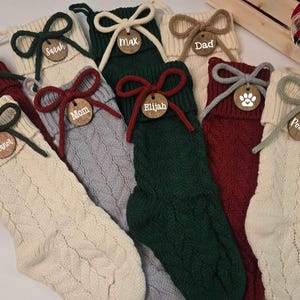May include: A selection of cable-knit Christmas stockings in cream, green, and red. Each stocking features a wooden tag with a name and a decorative bow. A festive holiday decoration, perfect for the Christmas season.