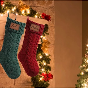 Peut inclure: Deux chaussettes de Noël en tricot avec un motif en tricot torsadé. La chaussette de gauche est turquoise et porte une étiquette indiquant "Mr.". La chaussette de droite est rouge et porte une étiquette indiquant "Mrs."