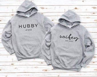 Peut inclure: Deux sweat-shirts à capuche gris avec du texte noir. Un sweat-shirt dit "Hubby est. 2022" et l'autre dit "wifey EST. 2023".
