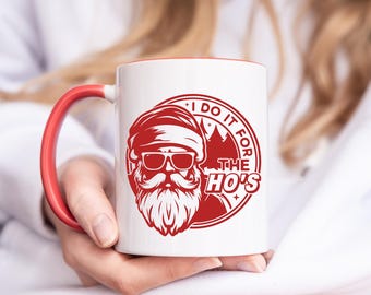 May include: White ceramic mug with a red interior and handle. The mug features a red graphic of Santa Claus wearing sunglasses and a beard, with the text "I DO IT FOR THE HO'S" in a circular design. A fun, novelty item.