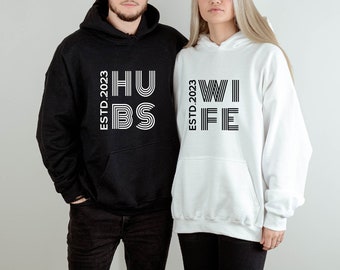 Peut inclure: Un sweat-shirt à capuche noir avec le texte "ESTD. 2023 HUSB" en blanc et un sweat-shirt à capuche blanc avec le texte "ESTD. 2023 WIFE" en noir.