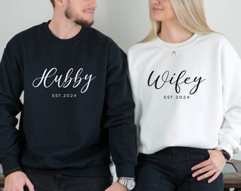 Peut inclure: Deux sweat-shirts assortis, un noir et un blanc. Le sweat-shirt noir porte l'inscription "Hubby" avec "EST. 2024" en dessous. Le sweat-shirt blanc porte l'inscription "Wifey" avec "EST. 2024" en dessous. Les sweat-shirts sont de style décontracté.