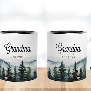 May include: Two white ceramic mugs with black rims. The mug on the left says "Grandma EST 2023" and the mug on the right says "Grandpa EST 2023". Both mugs feature a watercolour design of a mountain range with pine trees.
