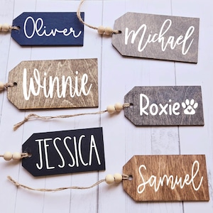 May include: Six wooden gift tags with names written in white paint. The tags are tied with twine and have wooden beads. The names on the tags are Oliver, Michael, Winnie, Roxie, Jessica, and Samuel.
