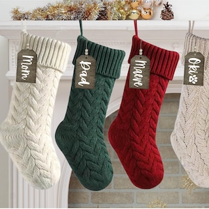 May include: Four knitted Christmas stockings with personalised wooden tags. The stockings are white, green, red, and white. The tags read "Mom", "Dad", "Maeve", and "Oki".
