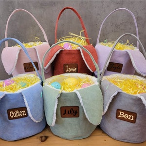May include: Five pastel-coloured Easter baskets with bunny ears. The baskets are made of corduroy fabric and have wooden name tags with the names Colton, Lily, Jamie,  Merl, and Ben.