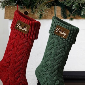 May include: Two cable-knit Christmas stockings, one red and one dark green, each featuring a folded cuff with a brown leather-like patch. The red stocking has the name Maddie engraved in white, and the green stocking has the name Wesley engraved.