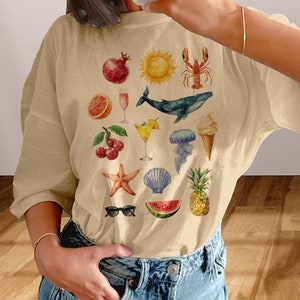 Può includere: Una maglietta beige con una varietà di illustrazioni a tema estivo, tra cui una anguria, un ananas, conchiglie, un'aragosta, una balena, una ciliegia, un pompelmo, un sole, un bicchiere da cocktail, occhiali da sole e una stella marina.