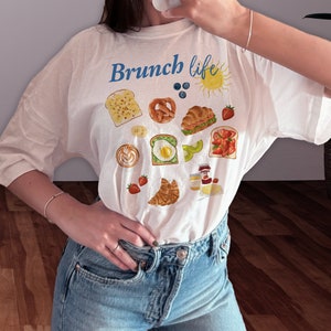 May include: White t-shirt with the text "Brunch life" in blue lettering and a variety of illustrated breakfast foods, including toast, bagels, croissants, fruit, and coffee.