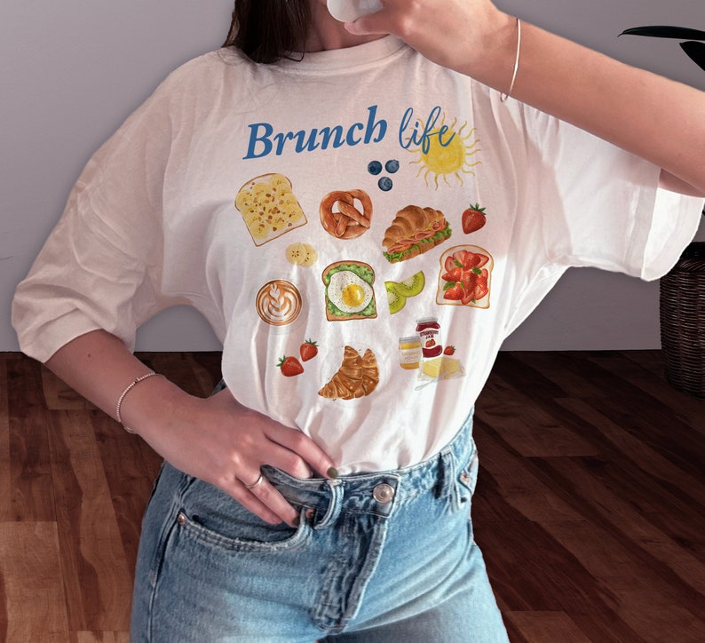 May include: White t-shirt with the text "Brunch life" in blue lettering and a variety of illustrated breakfast foods, including toast, bagels, croissants, fruit, and coffee.