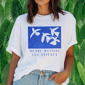 May include: White t-shirt featuring a blue rectangle with white bird silhouettes. The text "HENRI MATISSE LES OISEAUX" is printed below the image. The shirt is made of a soft material and has short sleeves.