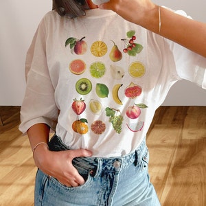 Cute girl wearing white T-shirt with print of mixed fruit: apple, orange, pear, cherries, pineapple, figs, citrus, kiwi, strawberry, pomegranate, lemon, lime, dragonfruit, grapes, peach, banana