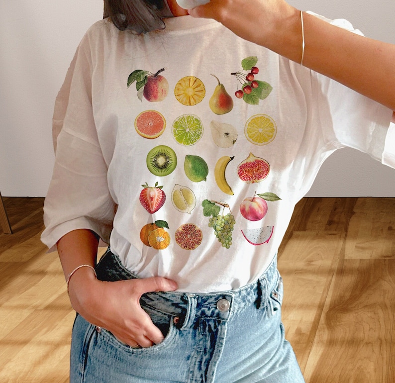 Cute girl wearing white T-shirt with print of mixed fruit: apple, orange, pear, cherries, pineapple, figs, citrus, kiwi, strawberry, pomegranate, lemon, lime, dragonfruit, grapes, peach, banana