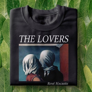 Pode incluir: Camiseta preta com uma estampa branca de uma pintura de René Magritte. A pintura retrata duas figuras com os rostos cobertos, se beijando. O texto "THE LOVERS" está acima da pintura.