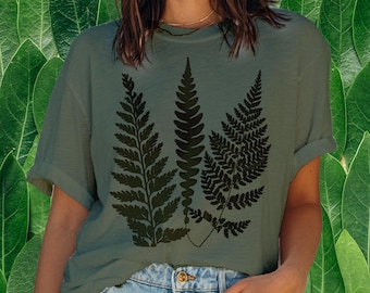 May include: An olive green t-shirt with a black fern leaf design. The short-sleeved top is made of a soft material and has a relaxed fit. The fern graphic is a detailed illustration of three different fern fronds.