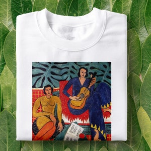 Puede incluir: Camiseta blanca con una pintura colorida de dos personas, una tocando la guitarra, sobre un fondo verde de hojas. La pintura es de estilo cubista con colores vivos y formas geom&eacute;tricas.