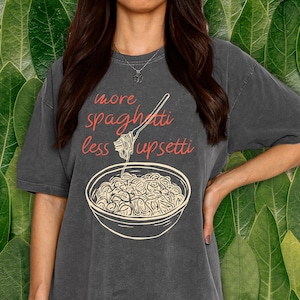 May include: Dark gray t-shirt with the text "more spaghetti less upsetti" in red script. A graphic of a bowl of spaghetti and a fork is below the text. The shirt is a casual style.