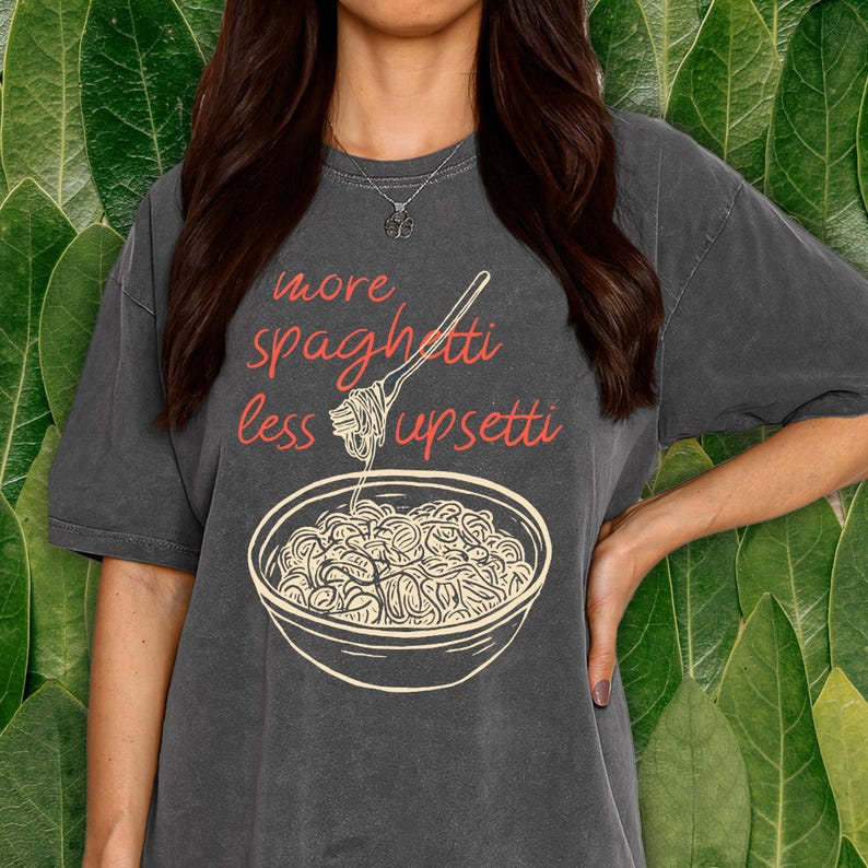 May include: Dark gray t-shirt with the text "more spaghetti less upsetti" in red script. A graphic of a bowl of spaghetti and a fork is below the text. The shirt is a casual style.
