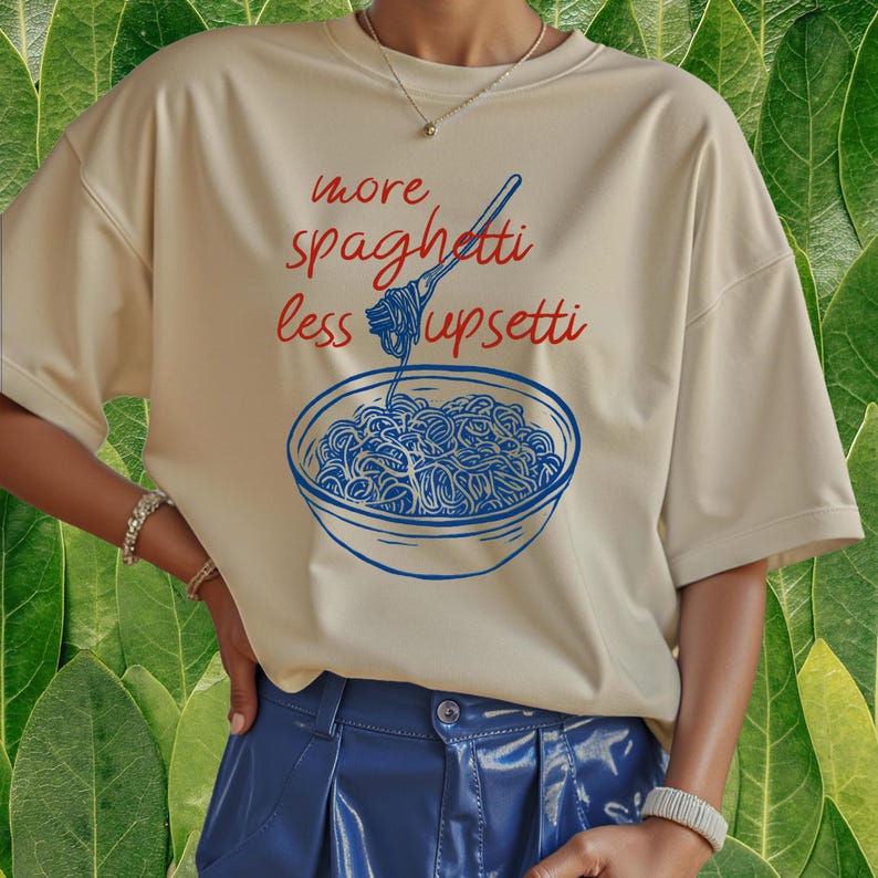 Puede incluir: Camiseta color crema con el texto "more spaghetti less upsetti" en rojo y una ilustraci&oacute;n azul de un plato de espaguetis. La camiseta tiene mangas cortas y un corte holgado.