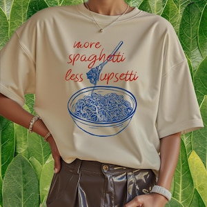 Puede incluir: Camiseta color crema con un gr&aacute;fico azul y rojo de un plato de espaguetis y el texto "more spaghetti less upsetti". La camiseta tiene mangas cortas y un corte holgado.