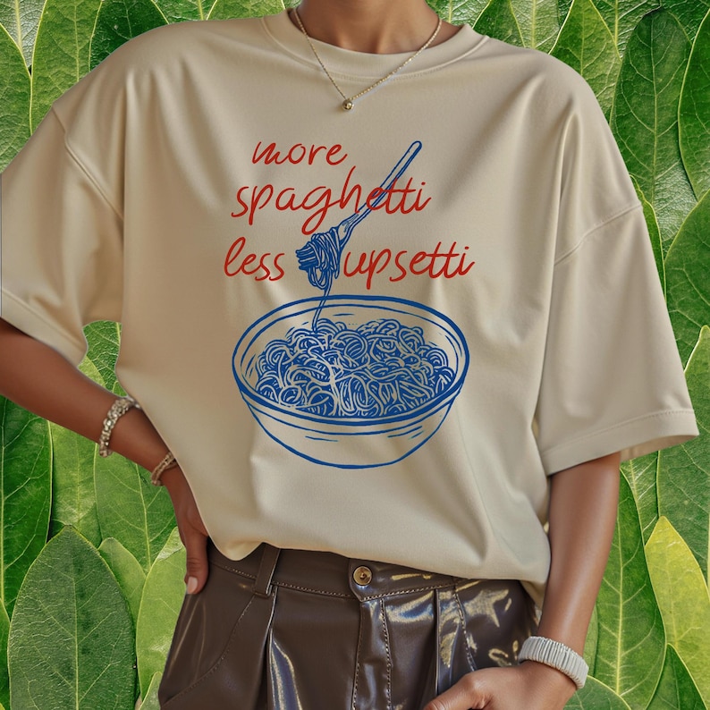 Puede incluir: Camiseta color crema con un gr&aacute;fico azul y rojo de un plato de espaguetis y el texto "more spaghetti less upsetti". La camiseta tiene mangas cortas y un corte holgado.