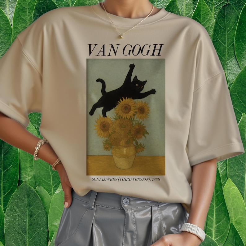 以下が含まれることがあります： ベージュ色のTシャツ。黒猫がゴッホの「ひまわり」のパロディであるひまわりの花瓶の上を飛び跳ねています。「VAN GOGH」の文字が画像の上にあり、「SUNFLOWERS (THIRD VERSION), 1888」が下にあります。
