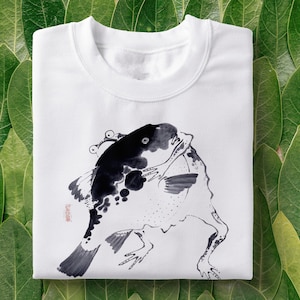 Pode incluir: Camiseta branca com uma ilustração em preto e branco de um peixe com um sapo nas costas.
