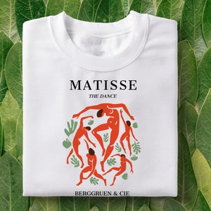 Pode incluir: Camiseta branca com uma ilustração vermelha e verde de dançarinos do quadro "A Dança" de Matisse. O texto "MATISSE THE DANCE" e "BERGGRUEN & CIE" está impresso na camiseta.