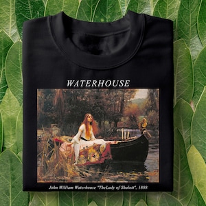 Pode incluir: Camiseta preta com uma pintura de uma mulher em um barco em um rio. A pintura é intitulada "The Lady of Shalott" de John William Waterhouse, 1888. WATERHOUSE está impresso em branco acima da pintura.