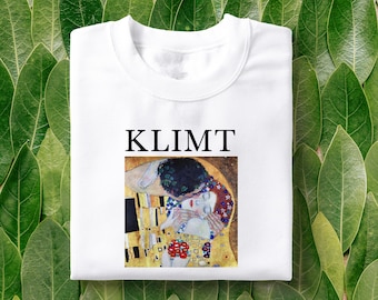 May include: White t-shirt featuring a black and gold print of Gustav Klimt's "The Kiss" painting. The text "KLIMT" is printed above the image.