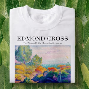 Pode incluir: Camisola branca com uma estampa colorida de uma paisagem costeira com duas mulheres. O texto "EDMOND CROSS" e "Two Women By the Shore, Mediterranean" são impressos acima da imagem. A camisola é exibida em folhas verdes.
